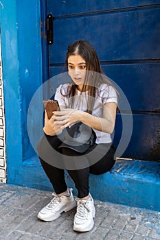 Portrait of a beautiful teenage girl checking messages on her cell phone sitting on the doorstep of a house. She looks surprised