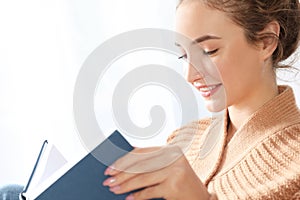 Portrait of beautiful teenage girl in warm cozy sweater reading book near window