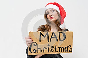 Portrait of a beautiful unhappy brunette woman in christmas hat holding a sign `Mad christmas ` on a white background. Vanity, hol