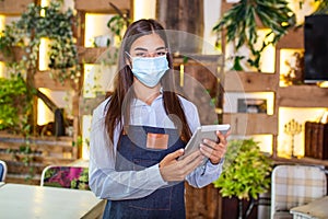 Portrait of beautiful waitress wearing protective face mask while holding touchpad and looking at the camera in a pub or