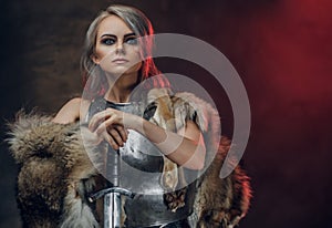 Portrait of a beautiful warrior woman holding a sword wearing steel cuirass and fur. Fantasy fashion. Studio photography