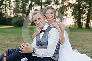 Portrait of beautiful wedding couple sitting on green grass in park in summer. Young cheerful woman bride hugging groom.