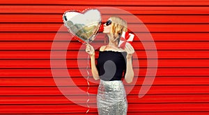 Portrait of beautiful woman blowing her lips sending sweet air kiss with gift box and silver heart shaped balloons on red