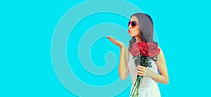 Portrait of beautiful woman with bouquet of red rose flowers and blowing her lips sending kiss in heart shaped sunglasses on blue