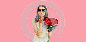 Portrait of beautiful woman with bouquet of red rose flowers and blowing her lips sending kiss in heart shaped sunglasses on pink