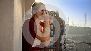 Portrait of beautiful woman drinking coffee at morning and looking on city from balcony. People relaxing, resting at home,