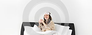 Portrait of beautiful woman enjoys her morning cup of coffee from bed, sitting in bedroom wearing sleeping mask and