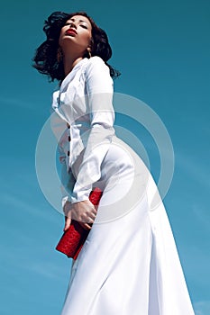 Portrait of a beautiful woman fashion model posing in elegant white atlas cocktail dress with red leather clutch in her hands