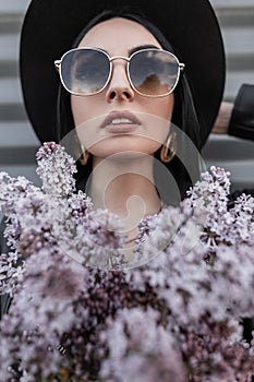 Portrait beautiful woman in fashionable sunglasses in chic elegant black hat with golden vintage earrings with lilac flowers near
