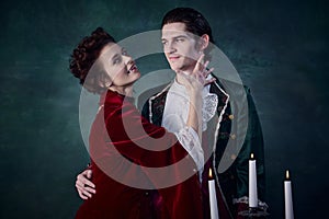 Portrait of beautiful woman with hypnotic eyes in image of vampire hugging young man, prince over dark green background.