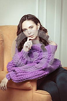 Portrait of beautiful woman with makeup, in knitted, purple oversize sweater, sitting on a chair on white background