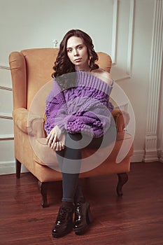 Portrait of beautiful woman with makeup, in knitted, purple oversize sweater, sitting on a chair on white background