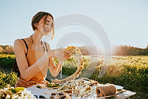 Portrait of beautiful woman making wreath of flowers dandelions on flowering field. Summer lifestyle, nature lover and freedom