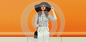 Portrait of beautiful woman model wearing black round straw summer hat, handbag and white striped shirt on orange background