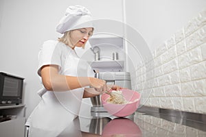 Portrait of beautiful woman pastry chef, wearing white hat and apron, mixing the flour in a pink bowl using a whisk