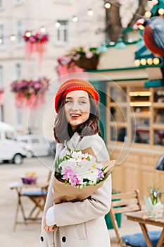 Portrait of a beautiful woman in a red beret and coat with a bouquet of flowers walks down the street