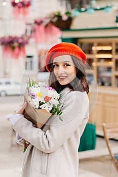 Portrait of a beautiful woman in a red beret and coat with a bouquet of flowers walks down the street