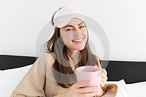 Portrait of beautiful woman relaxing in bed, waking up and having her morning cup of coffee in bedroom, looking happy