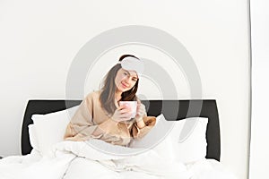 Portrait of beautiful woman relaxing in bed, waking up and having her morning cup of coffee in bedroom, looking happy