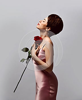 Portrait of beautiful woman with short hair in pink silk dress and earrings standing, holding red rose in hands with eyes closed