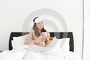 Portrait of beautiful woman in sleeping bag and pyjamas, drinking coffee in bed, enjoying her bright, relaxing morning