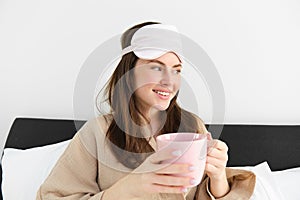 Portrait of beautiful woman in sleeping bag and pyjamas, drinking coffee in bed, enjoying her bright, relaxing morning