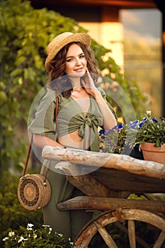 Portrait of beautiful woman smiling with curly hair in hat enjoying at park  by wooden cart with green grass and flowers. Sunset