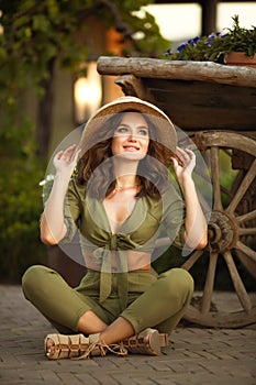 Portrait of beautiful woman smiling with hat enjoying at park. Attractive brunette with curly hair sitting by wooden cart with