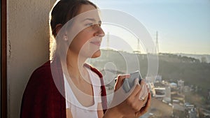 Portrait of beautiful woman standing on balcony with coffee and looking on sunrise at city. People relaxing, resting at home.