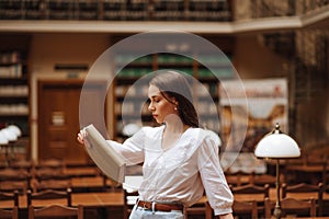 Portrait of a beautiful woman in a white blouse standing with a book in hand in a public library and reading a book with a serious