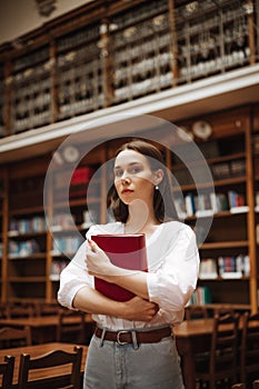 Portrait of a beautiful woman in a white blouse standing in a public library with a book in her hands and posing for the camera.