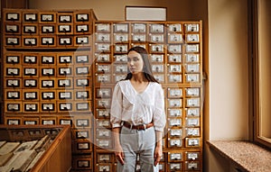 Portrait of a beautiful woman in a white blouse stands in the library archives on a background of boxes and looks away