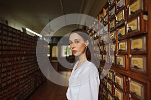 Portrait of a beautiful woman in a white blouse stands in the library archives and poses for the camera with a serious face