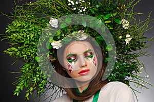 Portrait of a beautiful woman in a white dress with a green ribbon and with branches on her head. Creative makeup