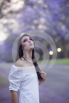 Portrait of beautiful woman in white dress standing in street surrounded by purple Jacaranda trees