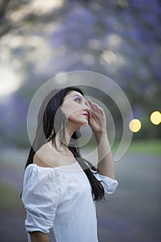 Portrait of beautiful woman in white dress standing in street surrounded by purple Jacaranda trees