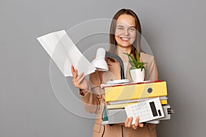 Portrait of beautiful young adult Caucasian woman wearing beige jacket posing isolated over gray background, holding paper folders