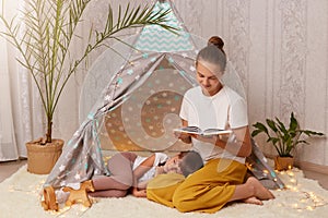 Portrait of beautiful young adult mother holding book and reading fairy tale for her little daughter, kid laying on floor and