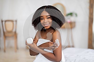 Portrait of beautiful young Afro woman enjoying morning skin care procedure, applying body cream at home