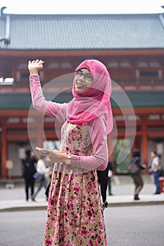 Portrait of beautiful young Asian muslim woman at gate of shrine in Japan waving hand and holding smart phone. Happy expression.