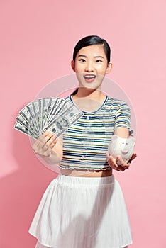 Portrait beautiful young asian woman with a lot of cash and money with piggy bank on pink isolated background