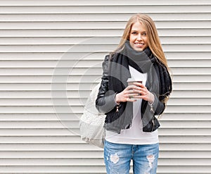 Portrait of a beautiful young blonde girl with long hair posing on a street with coffee and a backpack. Look at the camera. Outdoo