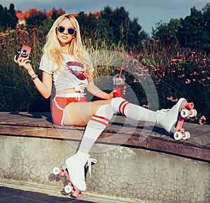 Portrait of a beautiful young blonde girl posing with a drink and a vintage red camera sitting on a bench in a vintage roller skat photo