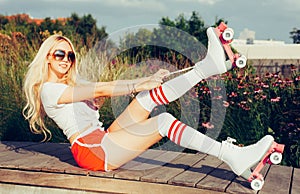 Portrait of a beautiful young blonde girl tighten the laces sitting on a bench in a vintage roller skates, wearing shorts, golfs a