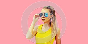 Portrait of beautiful young blonde woman model looking away wearing sunglasses, yellow t-shirt on pink studio background