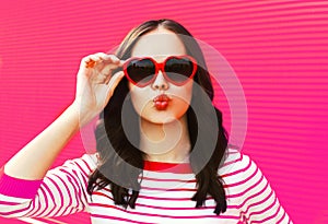 Portrait of beautiful young brunette woman blowing her lips wearing red heart shaped sunglasses on pink background
