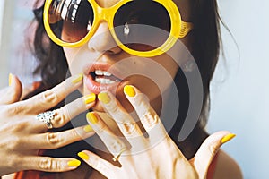 Portrait of a beautiful young brunette woman with full lips in yellow retro sunglasses and yellow varnish on the nails