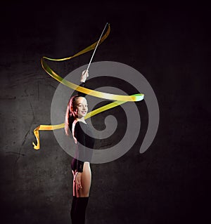 Portrait of beautiful young brunette woman gymnast training calisthenics exercise with yellow ribbon on dark studio background.