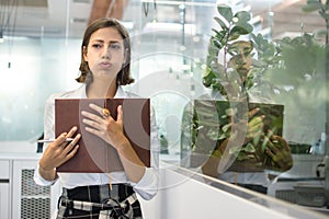 Portrait of beautiful young business woman pouting and holding notebook in office.