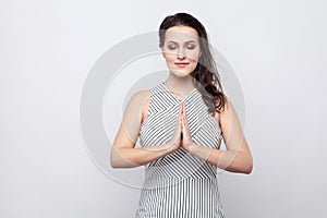 Portrait of beautiful young calm brunette woman with makeup and striped dress standing with closed eyes and palm hand in yoga pose